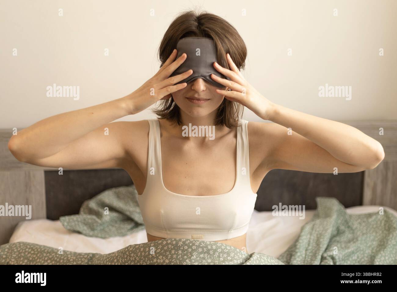 Eine junge Frau, die eine Schlafmaske auf dem Bett trägt und Hände nahe ihren Augen hält Stockfoto