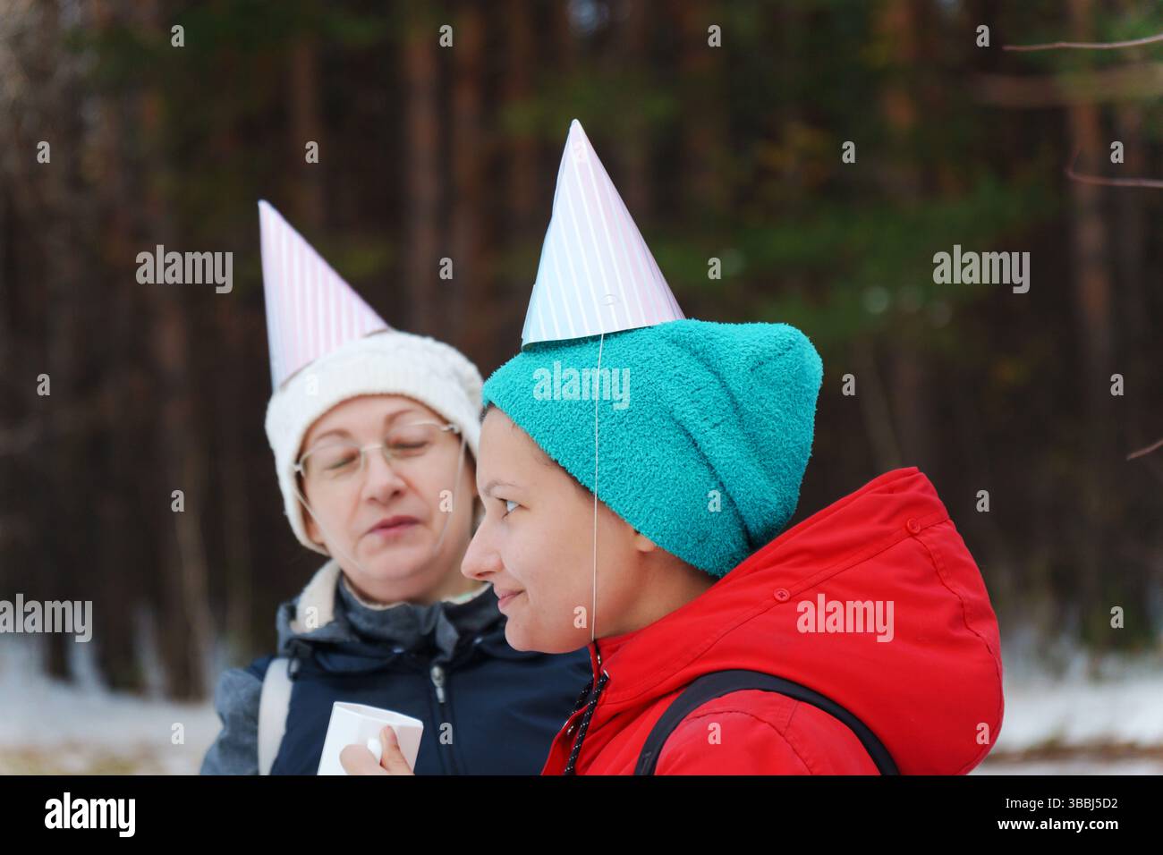 Zwei Freunde genießen im schneebedeckten Wald winterliche Leckereien und feiern mit festlichen Partyhüten Stockfoto