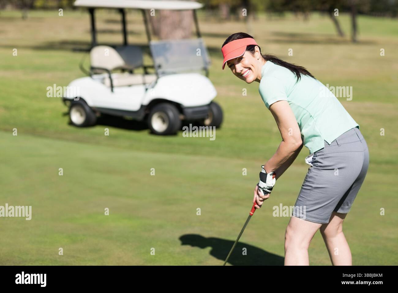 Lächelnde Frau mit Korallenvisier, die Putter in der Nähe von Golfball auf Putting Green hält, Kopierraum Stockfoto