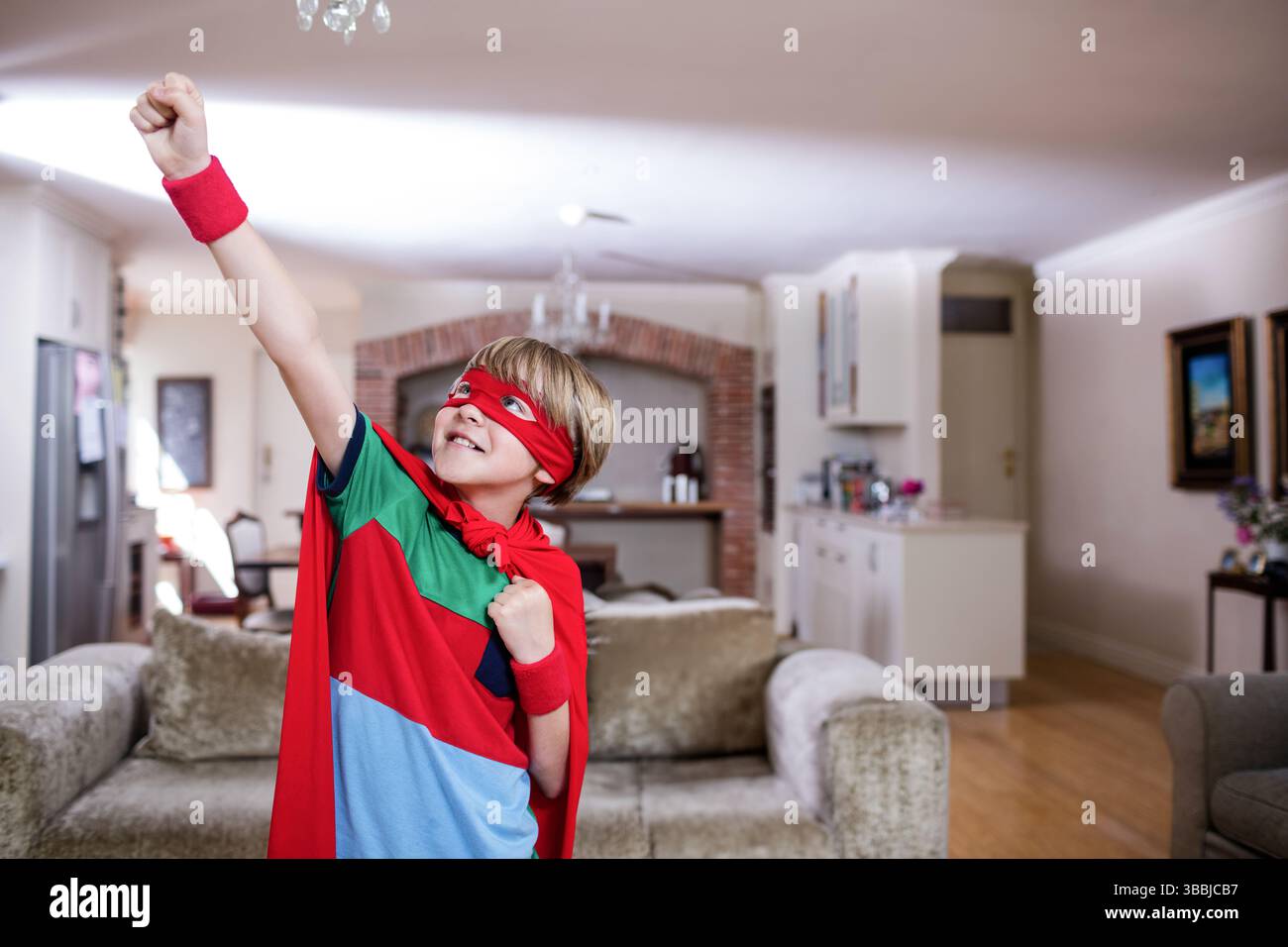 Blonder Junge steht im Wohnzimmer und hebt die Faust mit roter Maske, cape und Armbändern Stockfoto