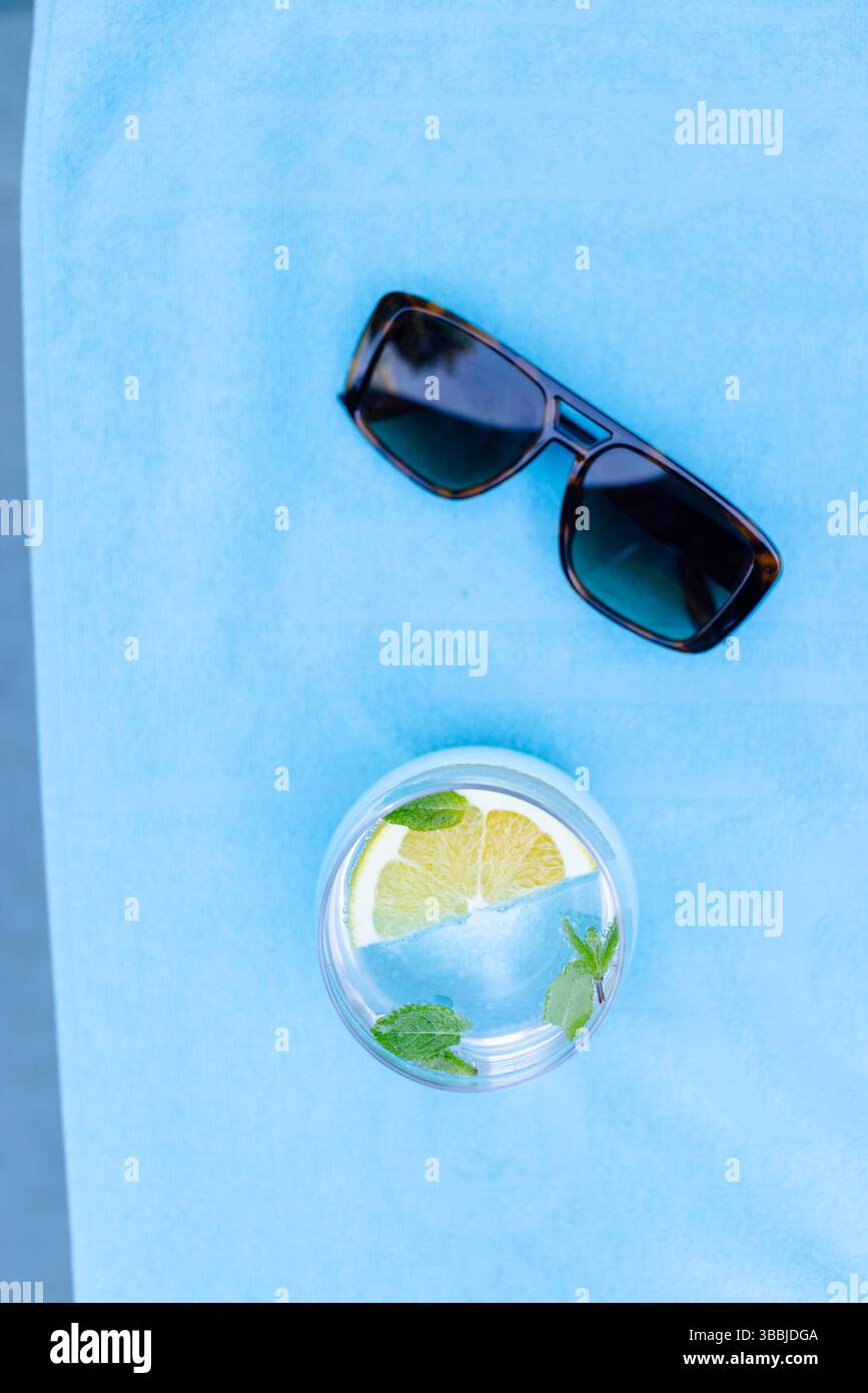 Glas Wasser mit Zitronenscheibe und Minzblättern auf blauem Handtuch, dunkle Sonnenbrille ruht Stockfoto