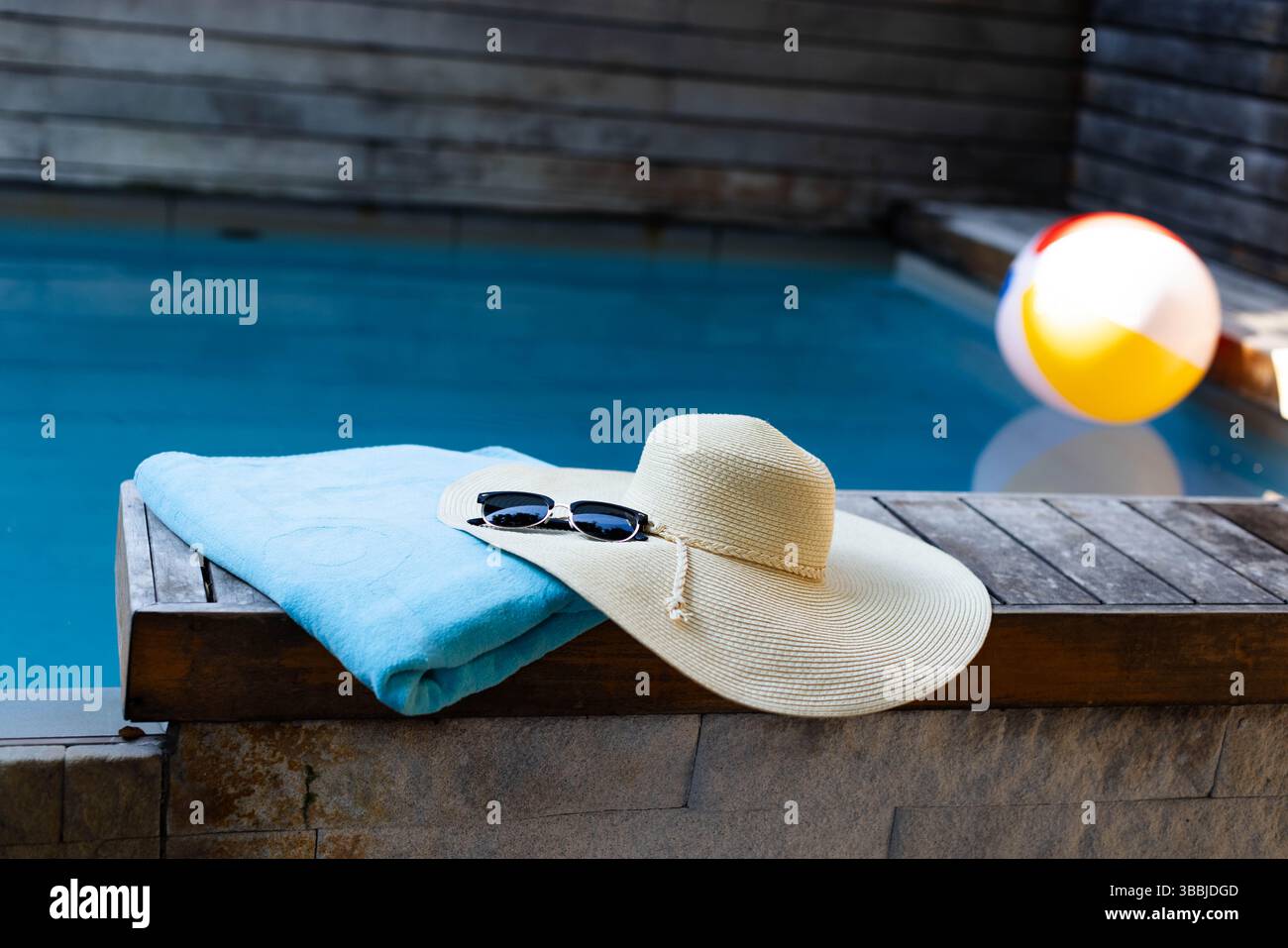 Holzbank hält Handtuch, Strohhut und Sonnenbrille in der Nähe des schwimmenden aufblasbaren Strandballs im Pool Stockfoto