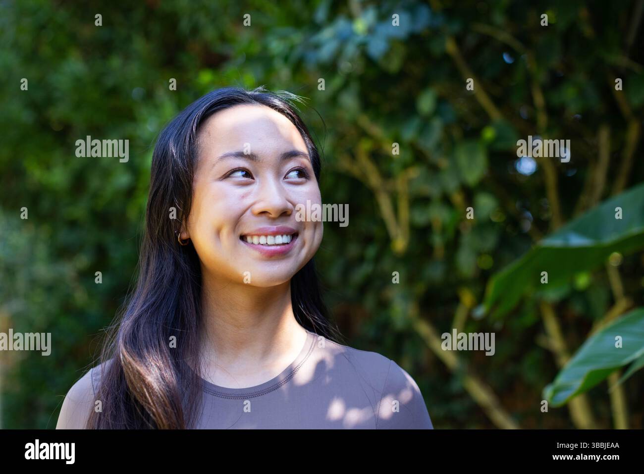 Lächelnde Chinesin mit malvenfarbenen Spitzen, die im Gartenlaub stehen und schimmerndes Sonnenlicht zeigen Stockfoto