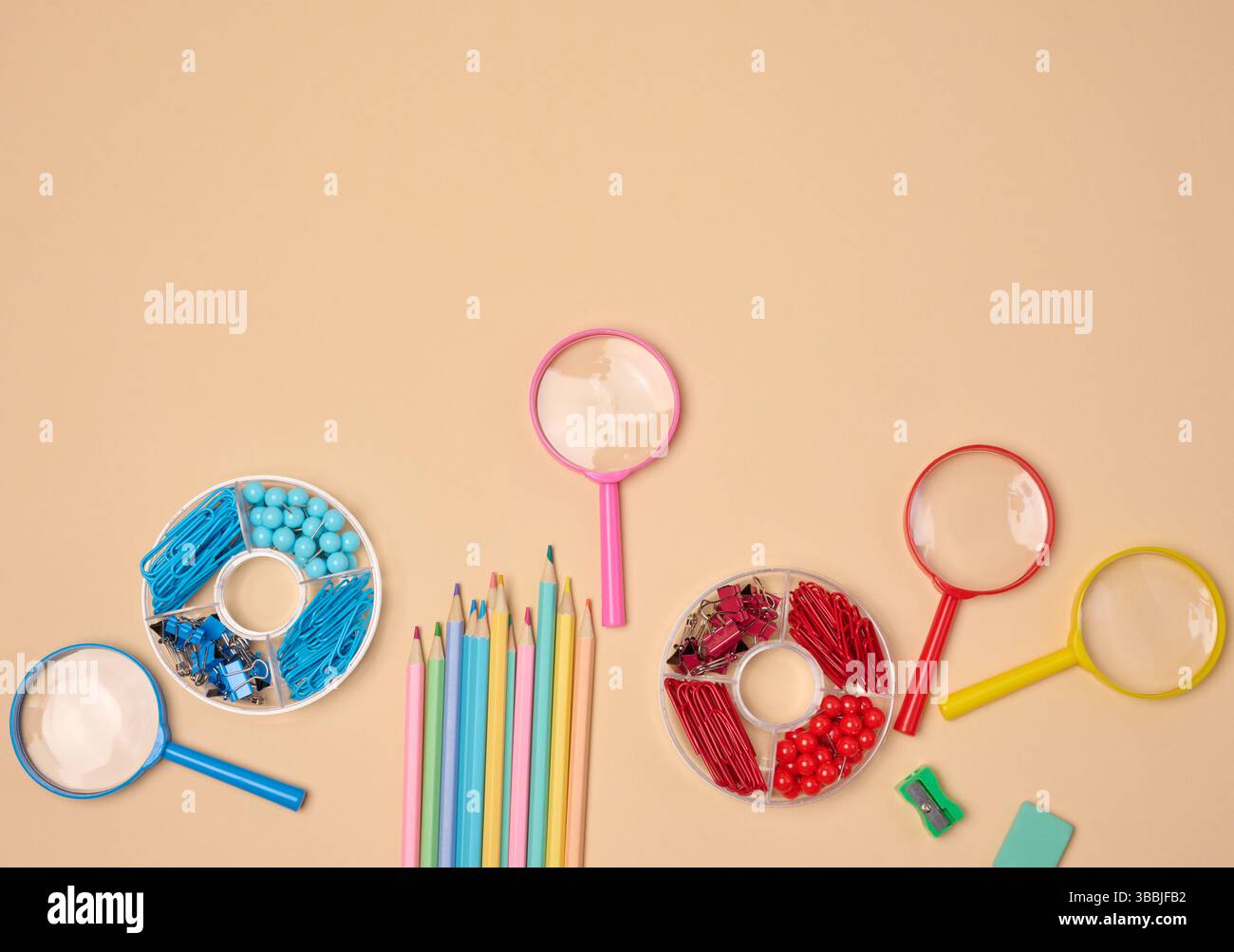 Verschiedene Büro- und Schulmaterialien, darunter Bleistifte, Lupen, Büroklammern und Pins, sind auf einem neutralen Hintergrund ordentlich angeordnet. Stockfoto