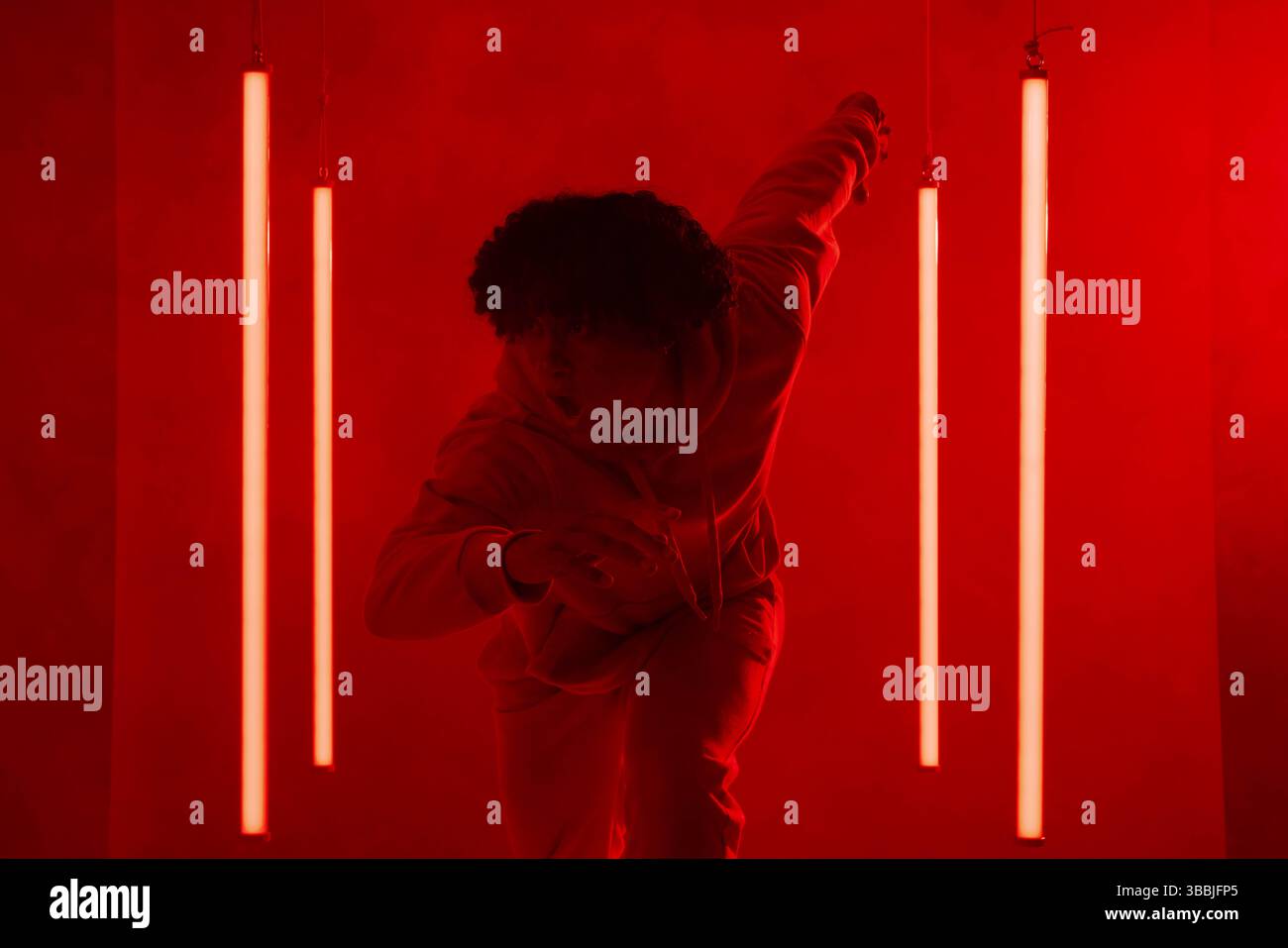 Ausbalancierter afroamerikanischer Mann im Hoodie, der Pose in rot beleuchtetem Studio mit LED-Röhrenlichtern zeigt Stockfoto