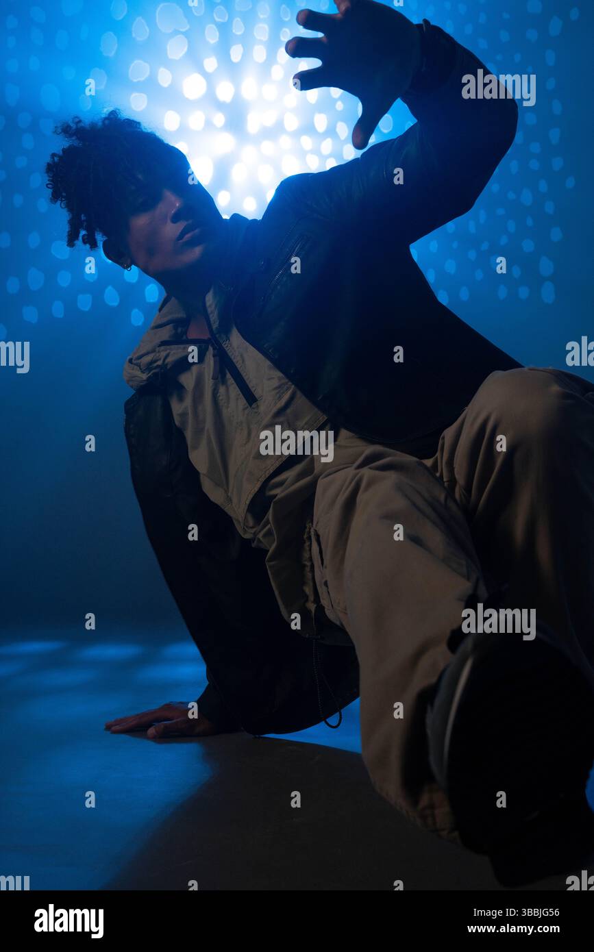 Tanzender Mann in Cargo-Hosen, Reißverschluss-Jacke in der Hocke auf der Bühne, mit blauem Raster Stockfoto