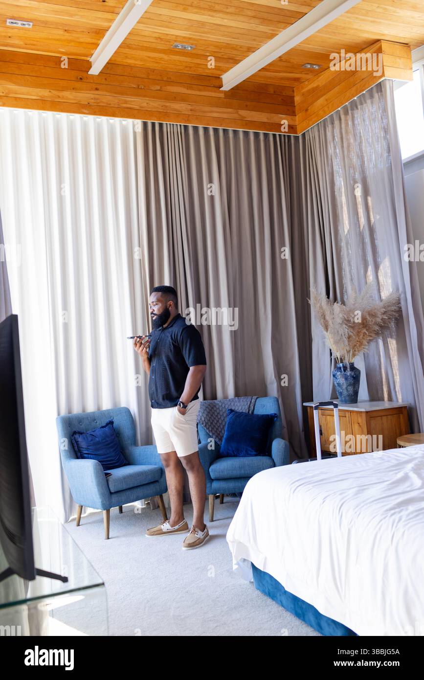 Sprechender Afroamerikaner, der in der Hotelsuite steht und das Smartphone in der Nähe von Sesseln hält Stockfoto Sprechender Afroamerikaner, der in der Hotelsuite steht und das Smartphone in der Nähe von Sesseln hält Stockfoto