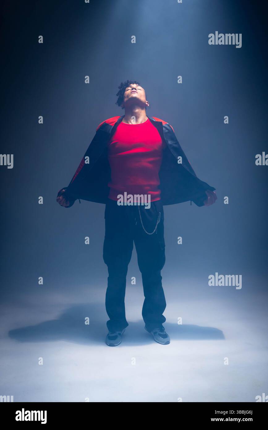 Posierender afroamerikanischer Mann mit rotem Oberteil und schwarzer Jacke mit Kette im Studio im Scheinwerferlicht Stockfoto