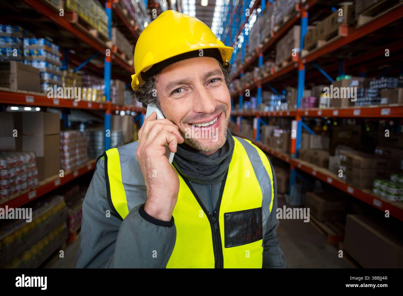 Das Smartphone am Ohr hält, reifer Erwachsener mit Sicherheitsweste, der im Lager mit Kisten spricht Stockfoto