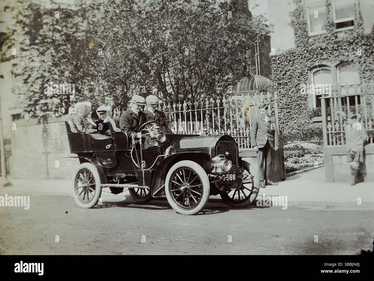 Ein altmodisches Auto aus dem frühen 20. Jahrhundert, 1907, mit vier Sitzplätzen. Das Fahrzeug hat ein unverwechselbares Vintage-Design und auf dem Kennzeichen steht „5988“. Es steht auf einer Straße vor einem in Efeu gehüllten Gebäude, was ihm einen zeitlosen Charme verleiht. Am Eingang befinden sich ein schmiedeeiserner Zaun und ein Tor, in dem zwei Personen in der Nähe stehen, die sich vielleicht unterhalten oder die Szene beobachten. Aus einem viktorianischen und edwardianischen Scrapbook, das scheinbar von einer unbenannten Person zusammengestellt wurde. Stockfoto