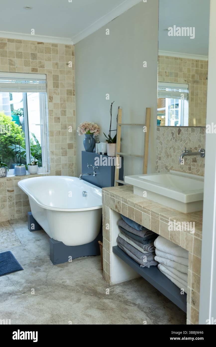 Die freistehende weiße Badewanne befindet sich im modernen Badezimmer mit einer Holzleiter mit rosafarbenen Blumen Stockfoto