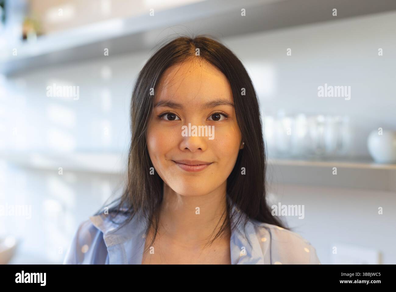 Posierende asiatische Frau mit Polka Dot-Shirt, die in der Küche steht, mit Metallregalen, Gläsern, Schüsseln Stockfoto