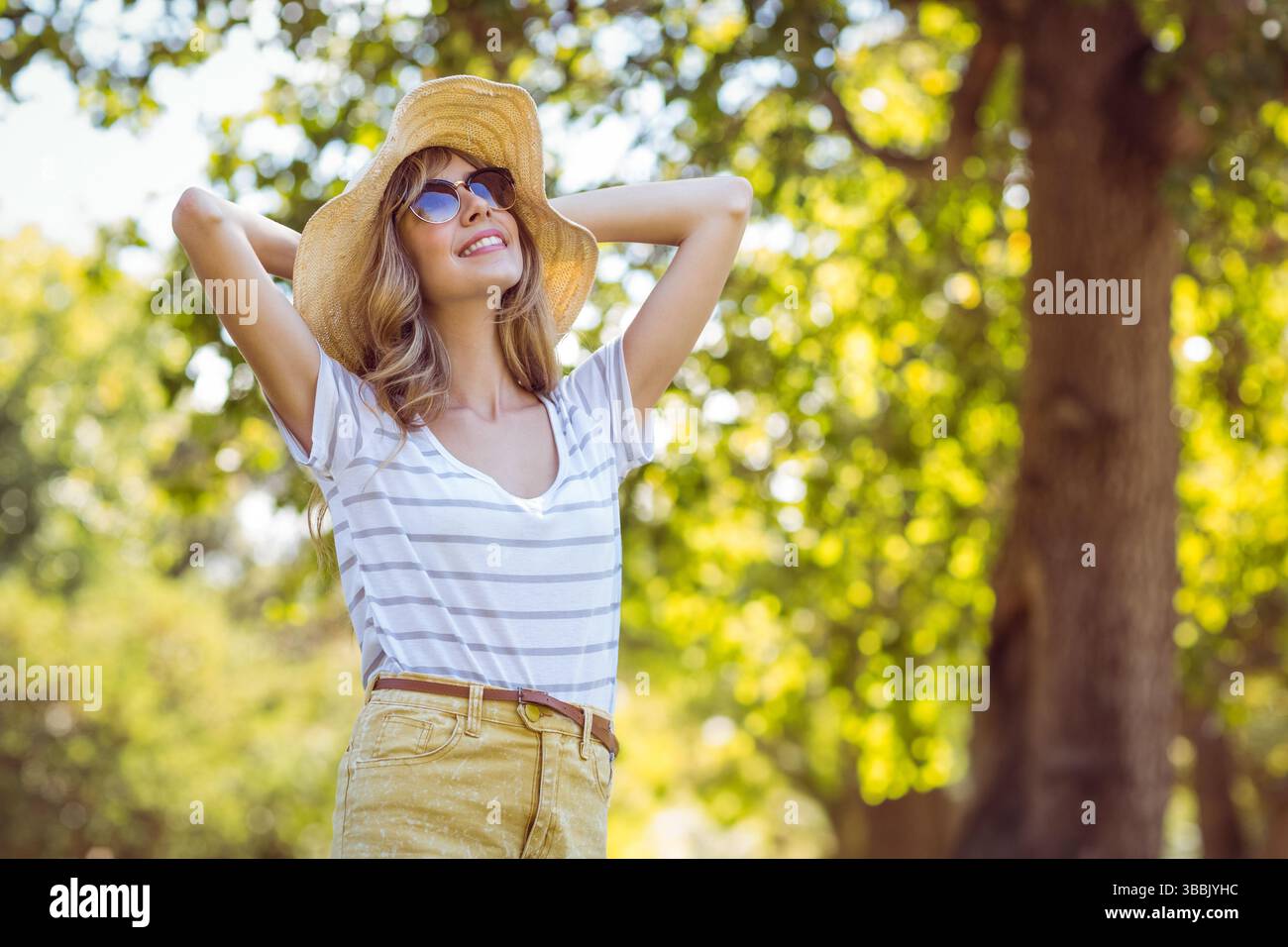 Frau, die Strohhut und Sonnenbrille unter dem Sonnenlicht im grünen Park in gelben Shorts justiert Stockfoto