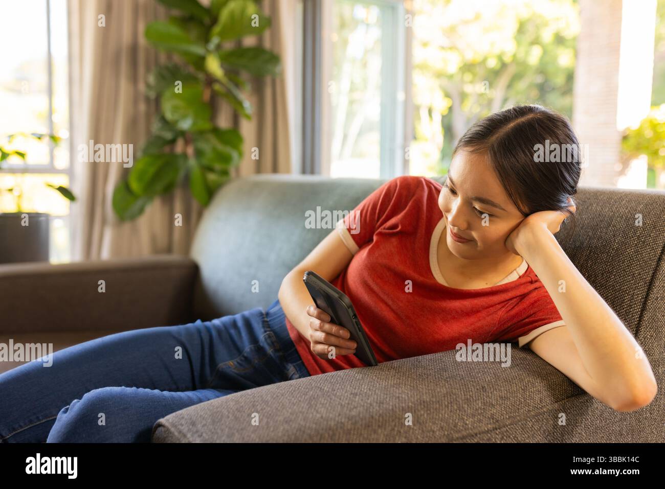 Asiatische Frau, die sich auf einem grauen Sofa im Wohnzimmer mit Topfpflanzen zurücklehnt, Smartphone hält, Kopierraum Stockfoto