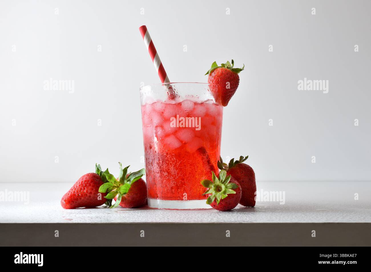 Erfrischendes Erdbeergetränk mit Eis in einer Glasschale, dekoriert mit einem Strohhalm und einer Scheibe Obst auf einem weißen Tisch mit Obst, Eis und Blättern mit einem Weißwein Stockfoto