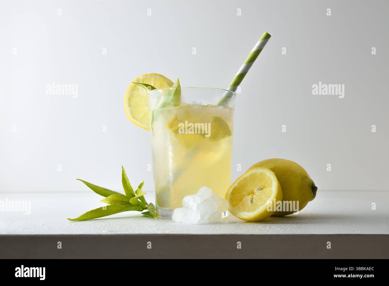 Erfrischendes Zitronengetränk mit Eis in einer Glasschale, dekoriert mit einem Strohhalm und einer Scheibe Obst auf einem weißen Tisch mit Obst, Eis und Blättern mit einem weißen IS Stockfoto