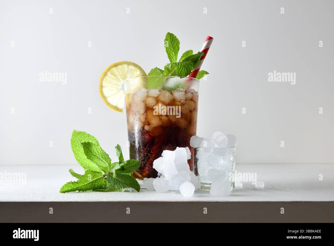 Erfrischendes Cola-Getränk mit Eis in einer Glasschale, dekoriert mit einem Strohhalm und einer Scheibe Obst auf einem weißen Tisch mit Obst, Eis und Blättern mit einem weißen iso Stockfoto