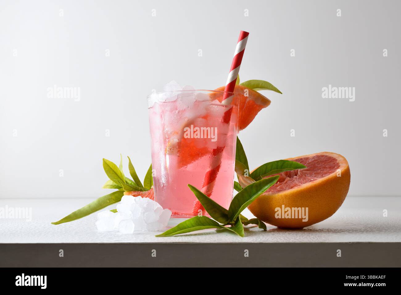 Erfrischendes Grapefruitgetränk mit Eis in einer Glasschale, dekoriert mit einem Strohhalm und einer Scheibe Obst auf einem weißen Tisch mit Obst, Eis und Blättern mit einem Whei Stockfoto