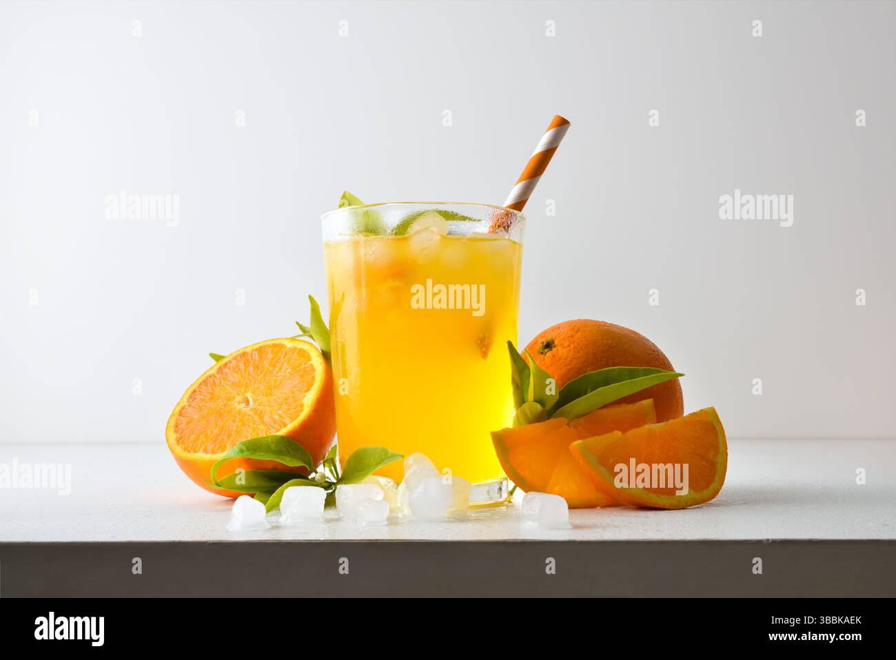 Erfrischendes Orangengetränk mit Eis in einer Glasschale, dekoriert mit einem Strohhalm und einer Scheibe Obst auf einem weißen Tisch mit Obst, Eis und Blättern mit einem weißen i Stockfoto