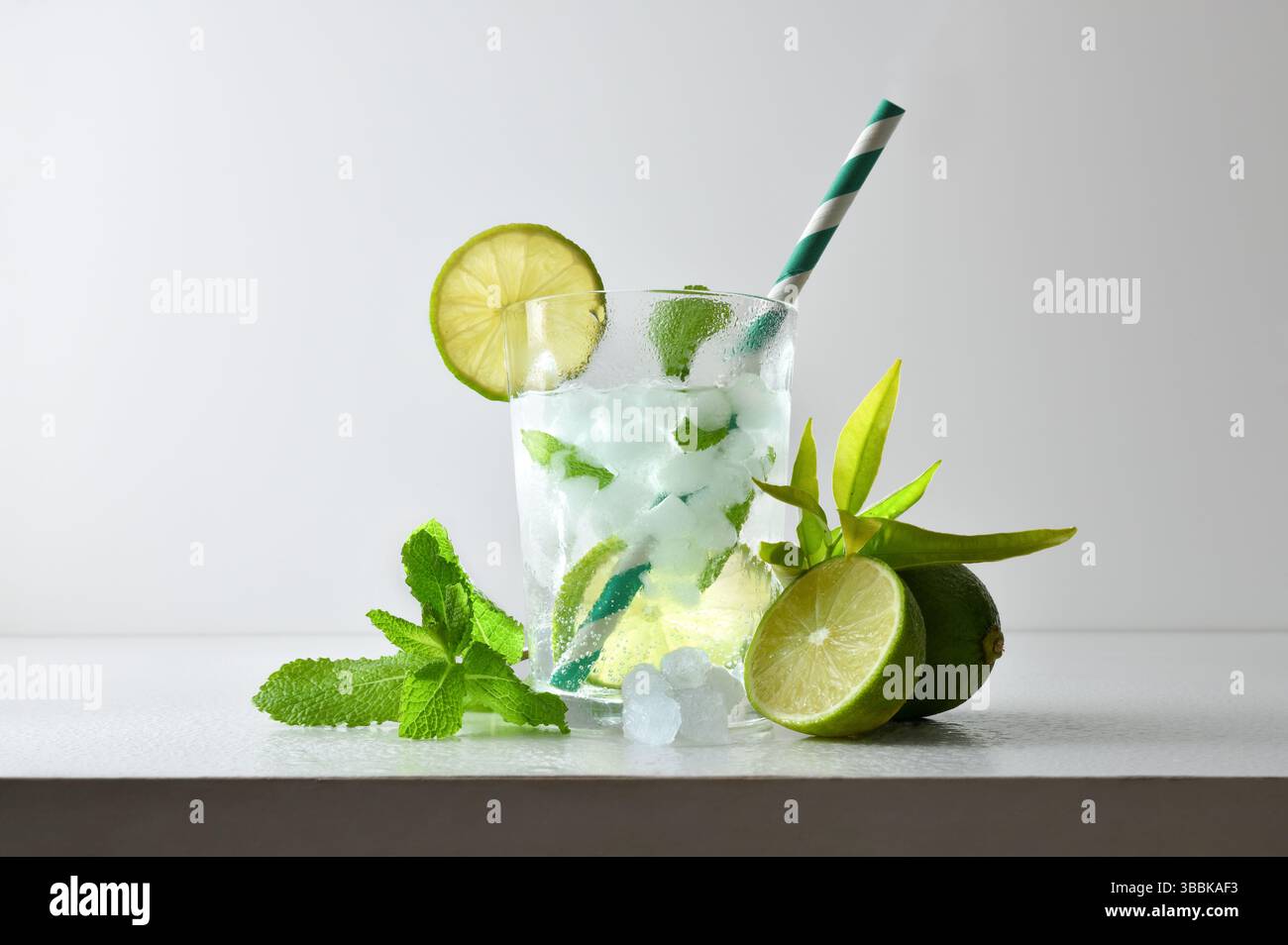 Erfrischendes Limetten- und Minzgetränk mit Eis in einer Glasschale, dekoriert mit einem Strohhalm und einer Scheibe Obst auf einem weißen Tisch mit Obst, Eis und Blättern mit A Stockfoto
