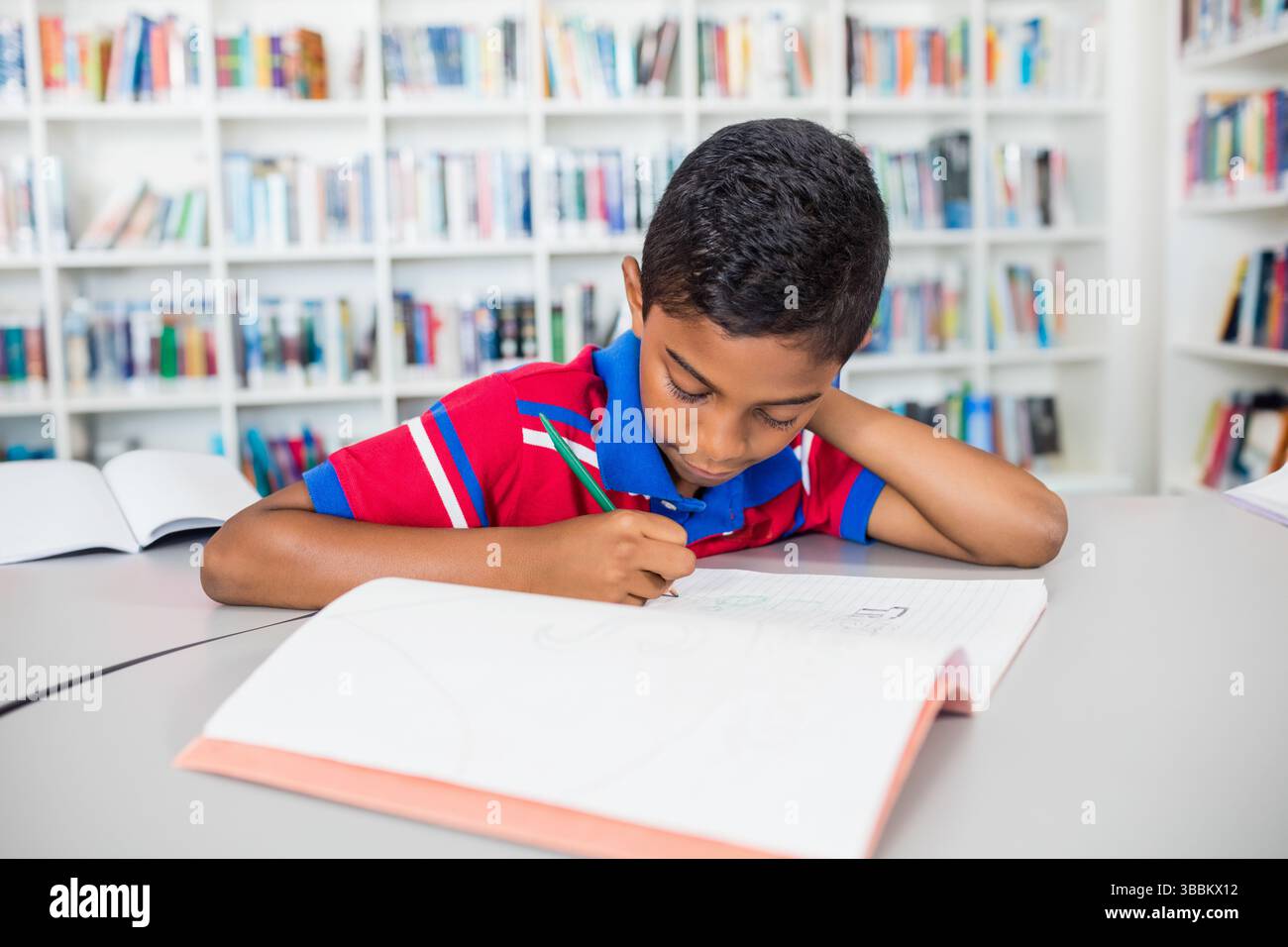 birassischer Junge, der in Rasternotizbuch an einem gekrümmten Bibliothekstisch schreibt, mit grünem Bleistift und offenen Büchern Stockfoto