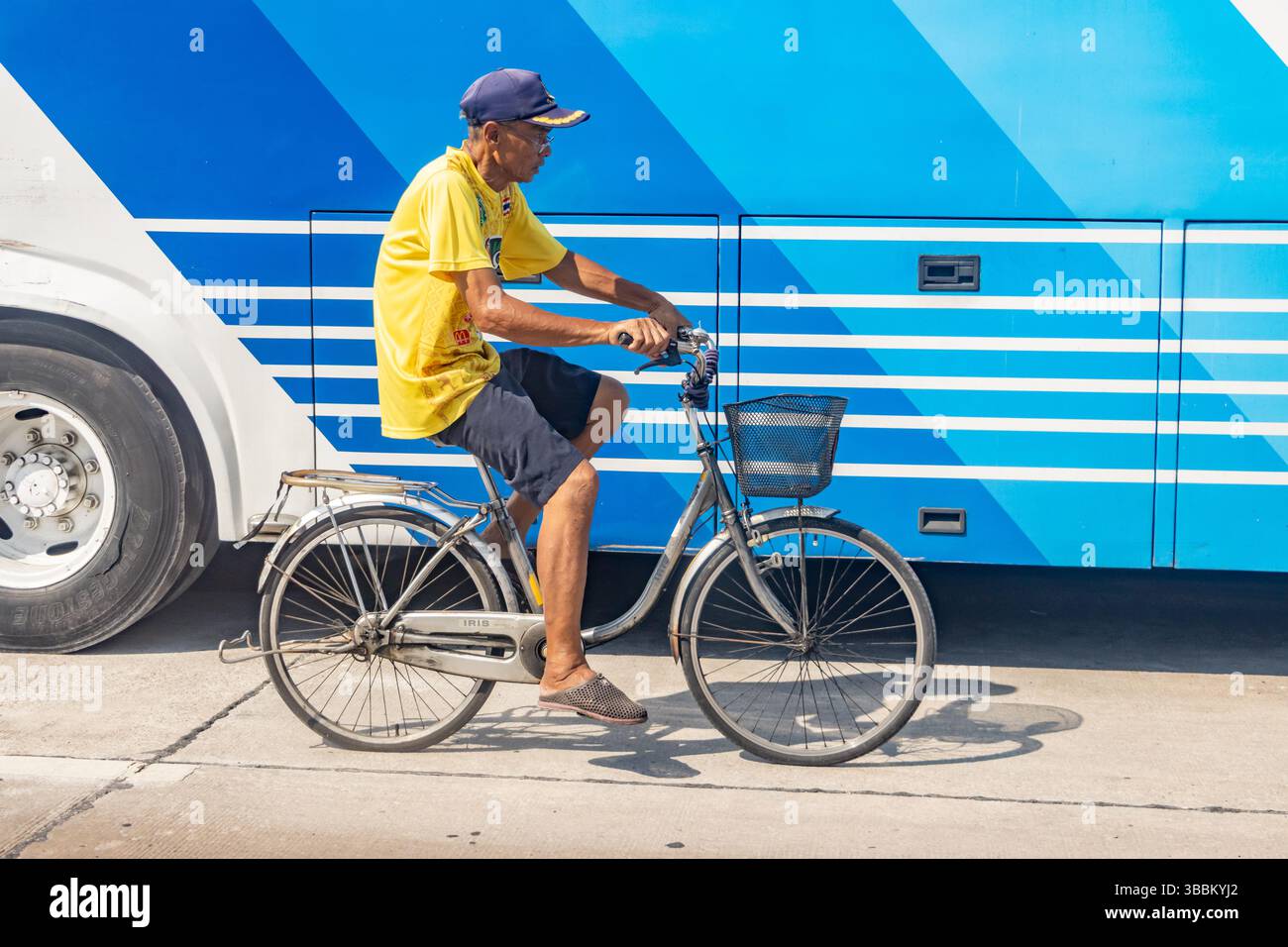 SAMUT PRAKAN, THAILAND, 28. April 2025, Mann auf dem Fahrrad auf einer Straße neben einem Bright Blue Bus Stockfoto
