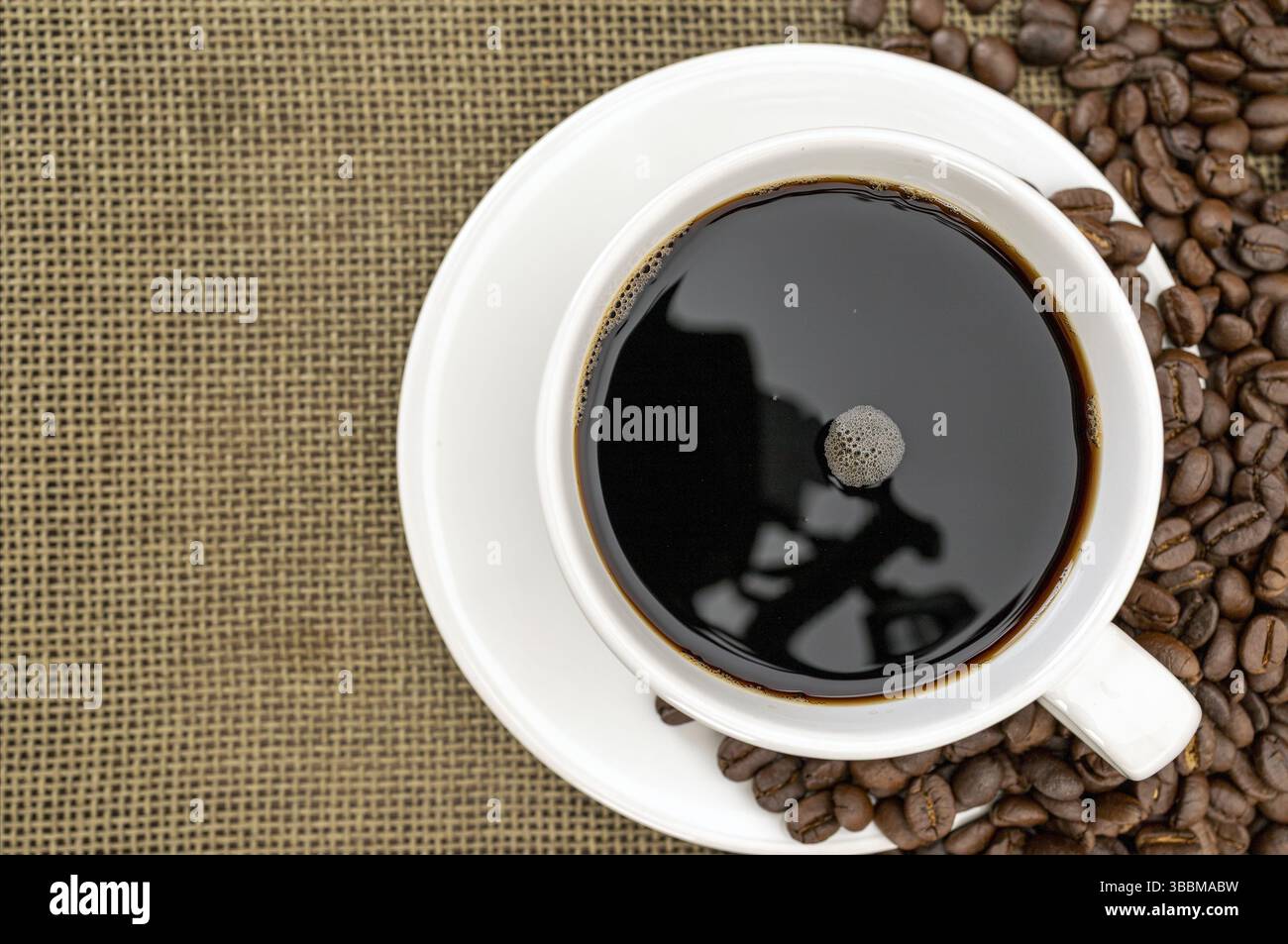 Eine dampfende Tasse schwarzen Kaffees sitzt auf einer weißen Untertasse, umgeben von verstreuten Kaffeebohnen auf der Oberfläche des Sackleines. Die Reflexion im Kaffee sorgt für einen Hauch von Intrige Stockfoto
