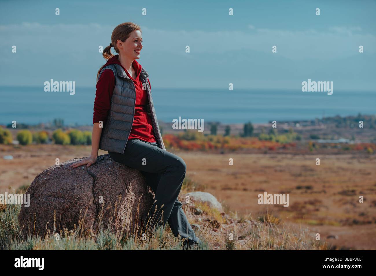 Junge lächelnde Touristin sitzt auf Stein auf See Hintergrund Stockfoto