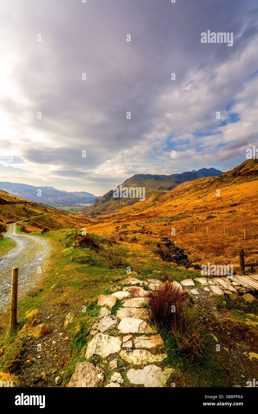 Schotterpfad mit Steinpfaden, der sich an heidekrauten Ufern vorbei zum Pen-y-Pass und den imposanten Hängen des Mount Snowdon unter einem dynamischen Himmel schlängelt Stockfoto