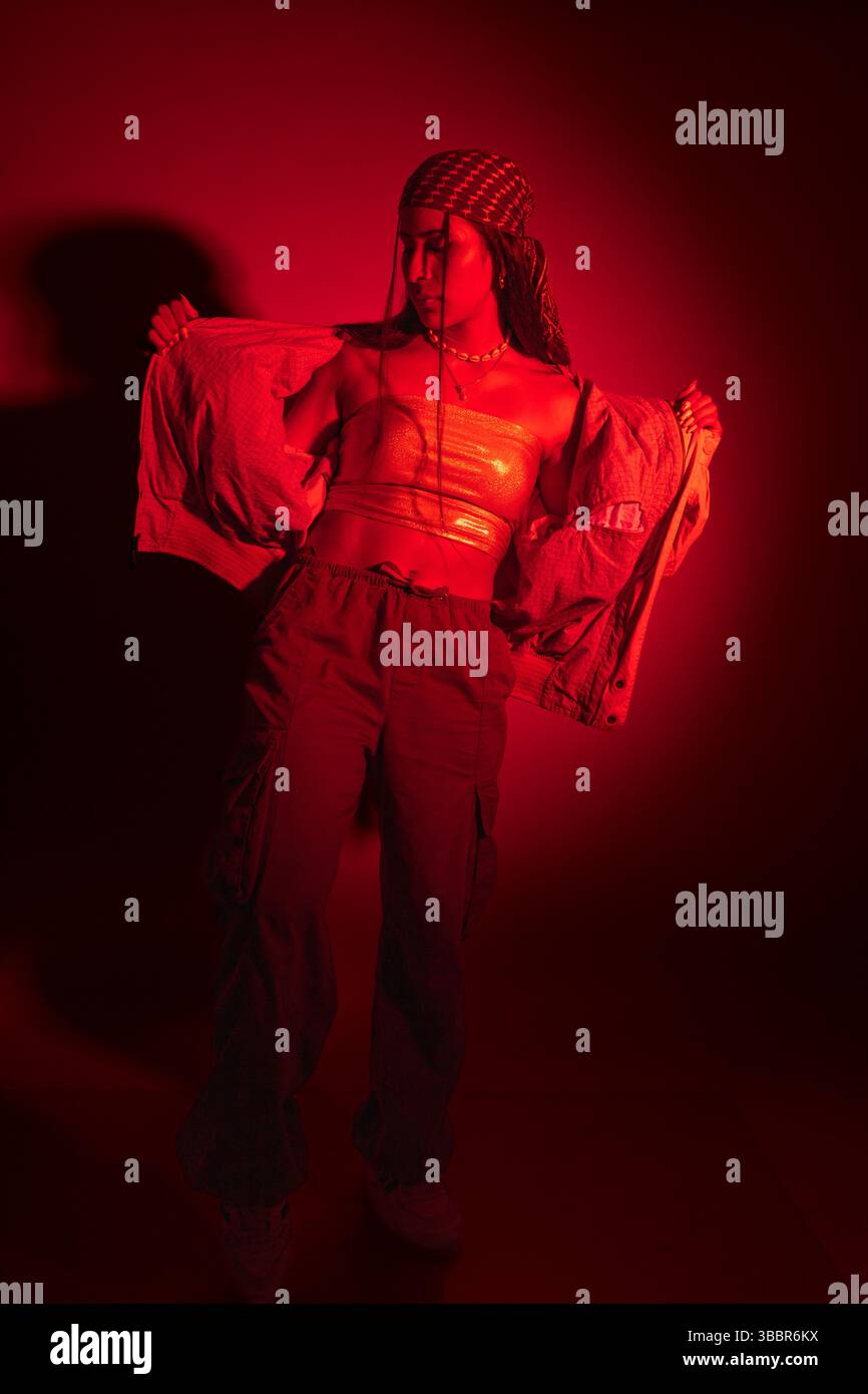 Eine junge Frau in roter Jacke und Hose posiert für ein Stadtfoto. Die rote Beleuchtung erzeugt eine dramatische und intensive Stimmung Stockfoto