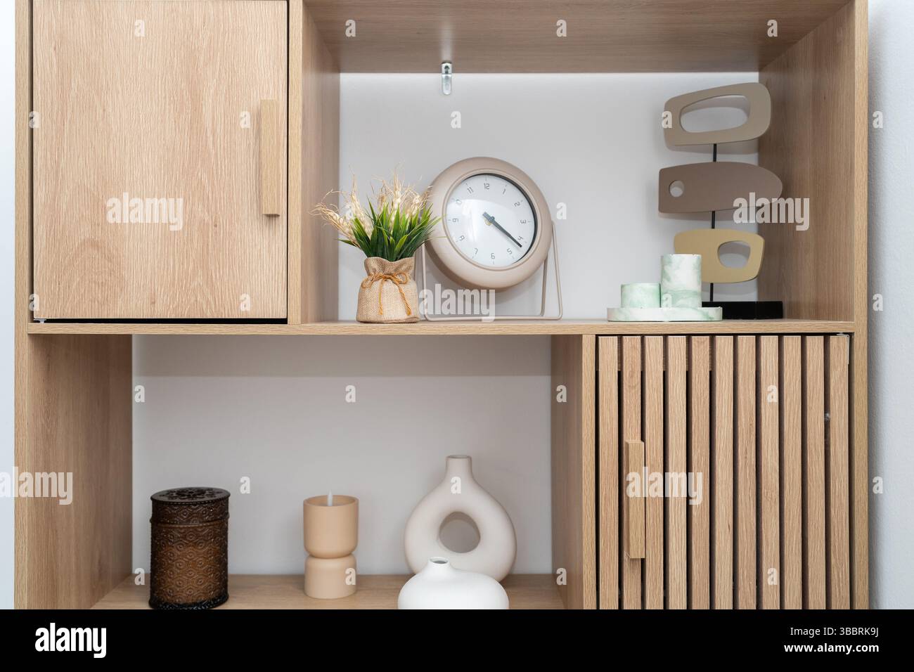 Ein gut organisiertes Holzregal bietet verschiedene moderne Dekorationsgegenstände, darunter eine stilvolle Uhr, Vasen und eine kleine Pflanze. Die Mischung aus Texturen verleiht dem Raum Wärme und Eleganz. Stockfoto