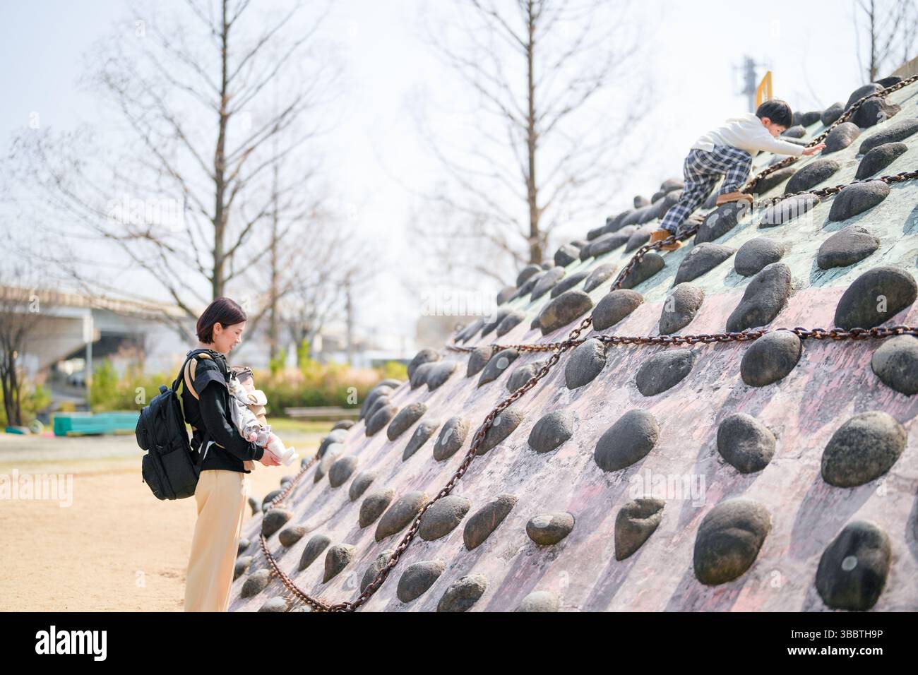 Im Frühjahr klettert ein dreijähriger Junge in einem Park in Fukuoka auf eine Felsstruktur. Seine Mutter und seine 6 Monate alte Schwester beobachten ihn. Dieses Foto zeigt eine warme und betrügerische Atmosphäre Stockfoto