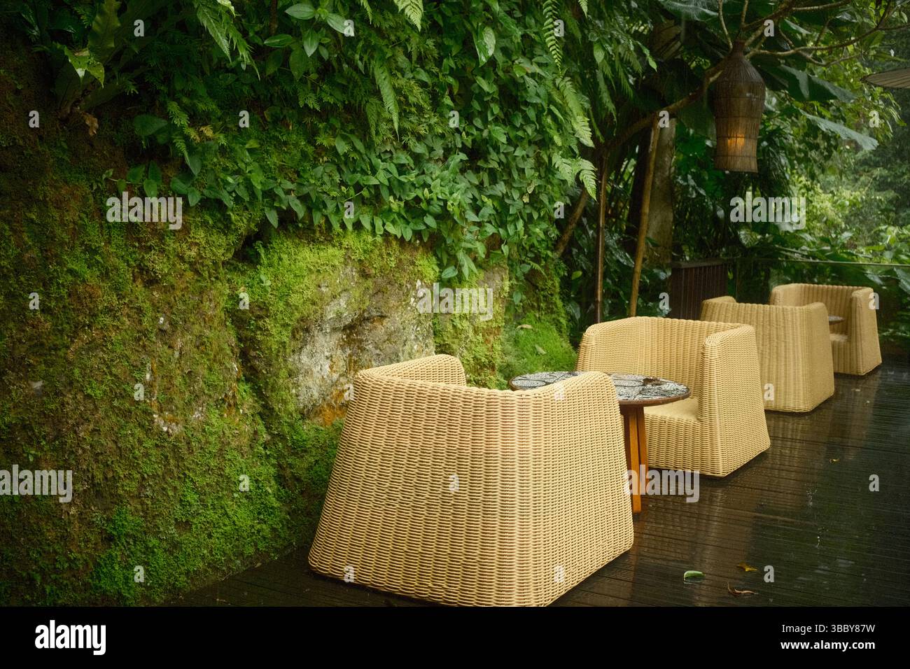Ruhige Sitzgelegenheiten im Freien mit gewebten Rattanstühlen, Holztischen und einer Holzterrasse neben einer moosigen Felsenwand Stockfoto