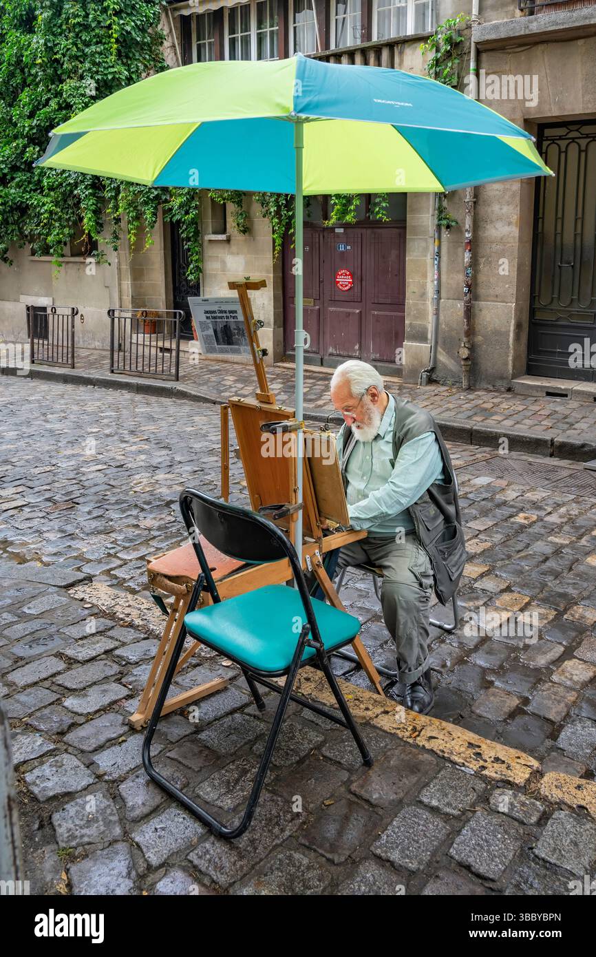 Paris, Frankreich, 12. Juli 2024: Ein alter Künstler mit weißem Bart sitzt auf einem Klappstuhl unter einem Sonnenschirm in einer kopfsteingepflasterten Straße in Montmartre. Stockfoto