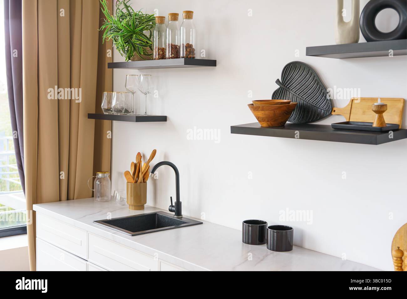 Weiße Küche mit Arbeitsplatte und schwarzem Wasserhahn auf weißem Wandhintergrund mit Holzregalen. Regale mit Utensilien, Küchenschrank und Einrichtung. Design in Stockfoto