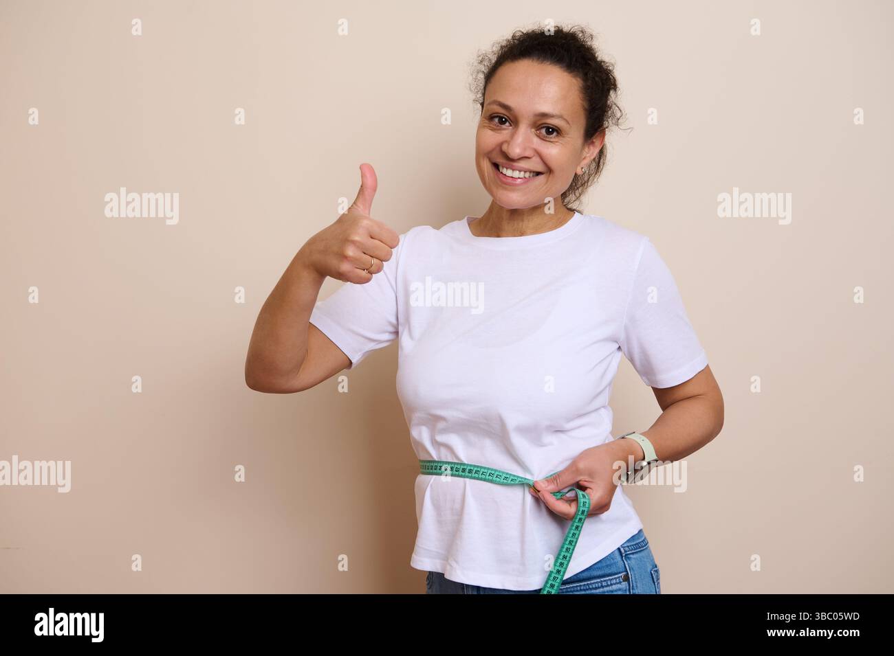 Die lächelnde Frau in lässigem weißem Hemd misst ihre Taille mit einem Klebeband und drückt ihre Begeisterung und Zufriedenheit über ihren Fortschritt beim Erreichen persönlicher fiktion aus Stockfoto