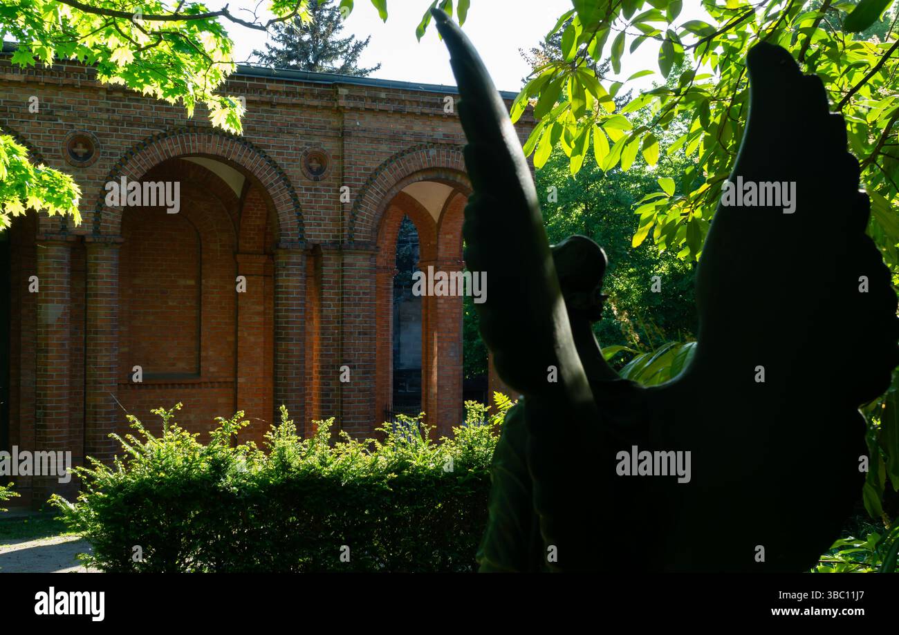 Silhouette einer Engelsstatue mit Blick auf die Backsteinkapelle auf dem Georgen-Pfarrfriedhof II in Berlin, umgeben von Sommergrün. Stockfoto
