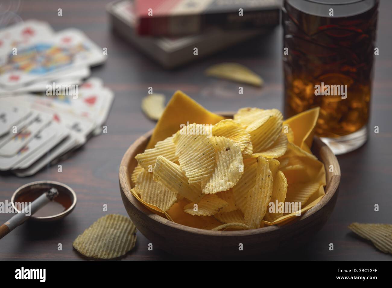 Schüssel mit Chips, Spielkarten und Zigarette neben einem Getränk auf einem Tisch Stockfoto