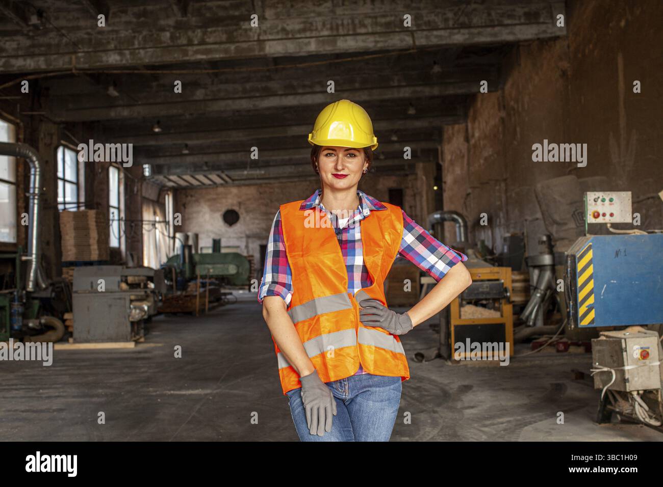 Eine wunderschöne junge Frau, die in der Möbelfabrik arbeitet und Jeans, eine orangefarbene Weste und einen gelben Schutzhelm trägt. Sie steht in der Holzwerkstatt und schaut auf die Stockfoto