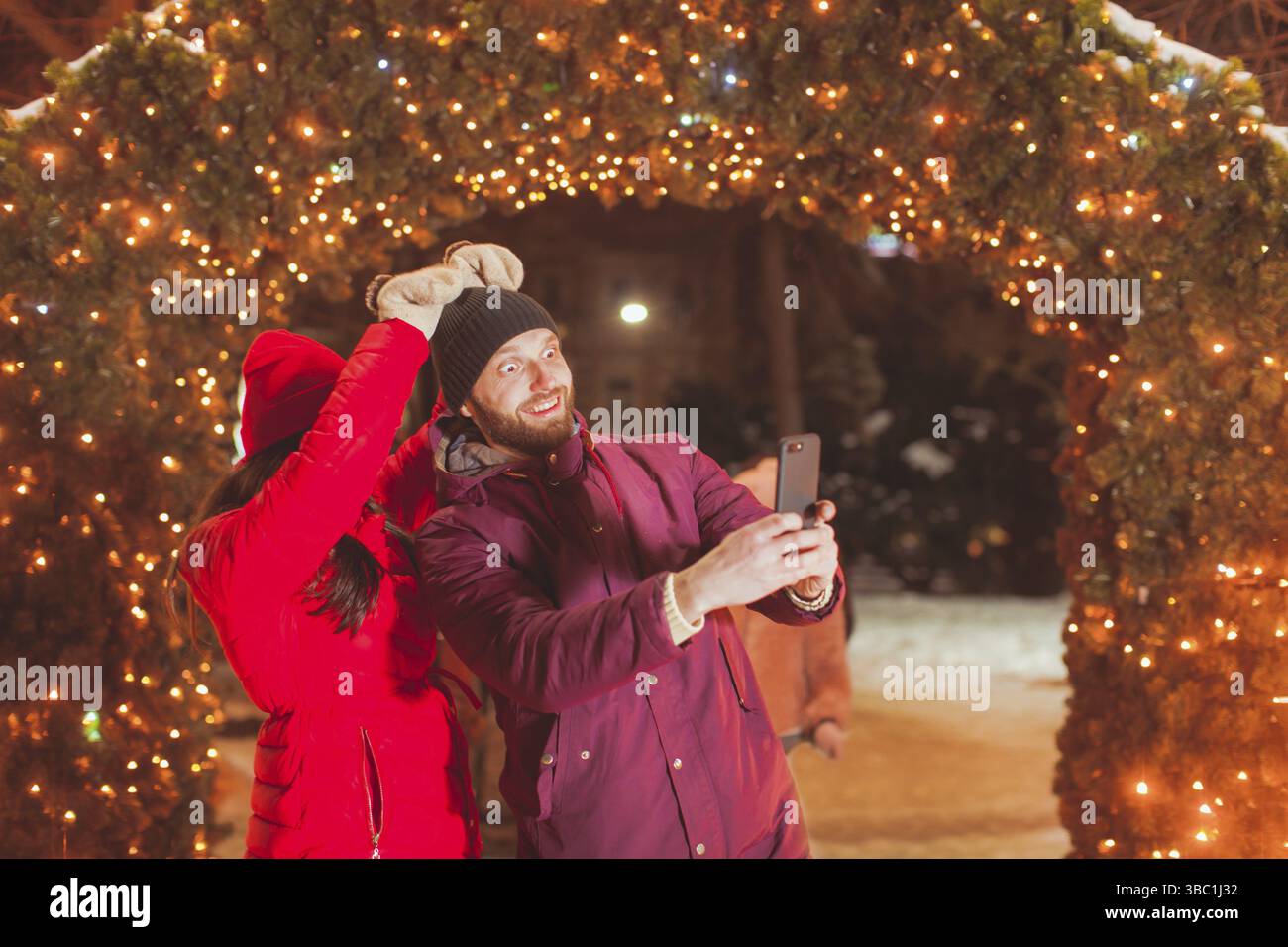 Junge glücklich, Mann und Frau, die selfie mit lustigen Gesichtern in der Nähe von Bogen mit Weihnachten Dekoration, Outdoor im Winter Markt Stockfoto