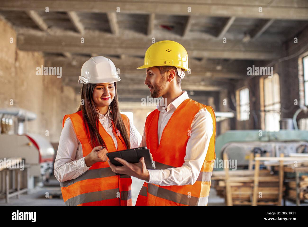 Lächelnde Arbeiter, die in der Fabrik arbeiten. Professionelle Helme, die im Lager ein Tablet benutzen Stockfoto