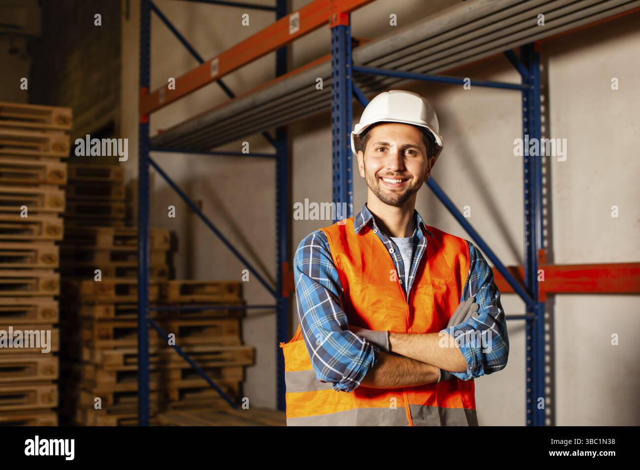 Selbstbewusster, gutaussehender Arbeiter in Schutzhelm und Uniform im Lager einer industriellen Fertigung. Vertrauen, Erfolg, Geschäftskonzept Stockfoto