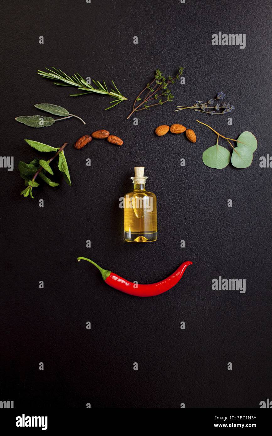 Lächelndes Gesicht aus Kräutern und einer Flasche ätherisches Öl auf schwarzem Hintergrund, Draufsicht, flach liegend. Gesunde organische, handgemachte Kosmetik Stockfoto