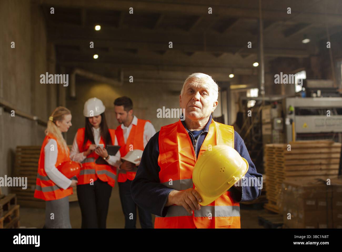 Porträt eines alten erfahrenen Fabrikarbeiters mit seinem Schutzhelm in den Händen, der im Geschäft der Holzfabrik steht. Gruppe junger Fachärzte für Schutzkleidung Stockfoto