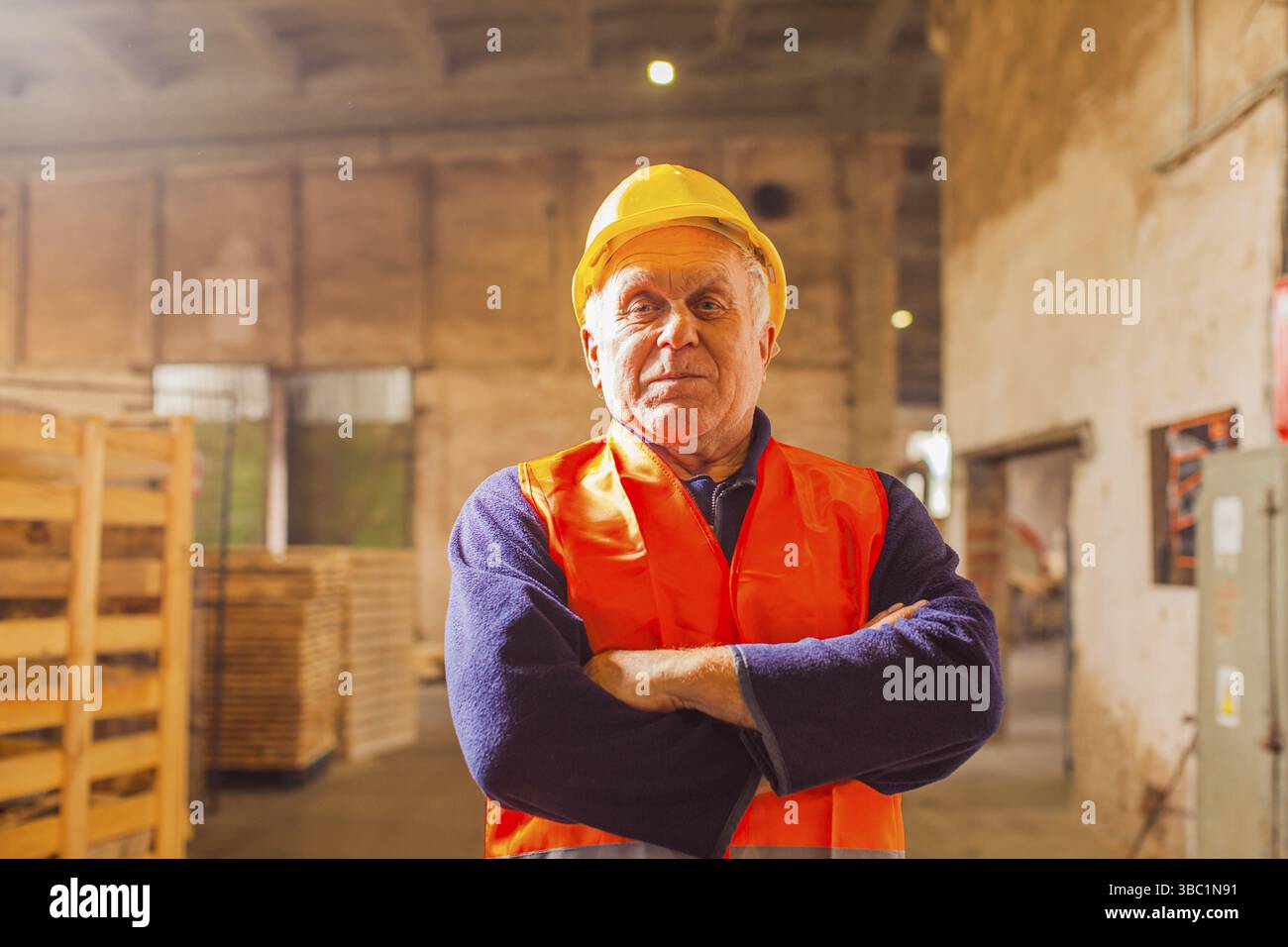 Porträt eines alten Mannes, selbstbewusster professioneller Arbeiter mit Bademantel, orangefarbener Weste und Helm, stehend in einer leeren Tischlerei Stockfoto