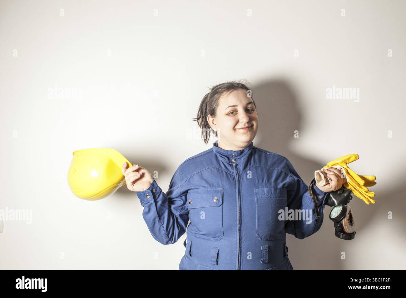 Lächelnde Arbeitnehmerin gekleidet in Arbeitskleidung, hält einen gelben Helm und gelbe Handschuhe Stockfoto