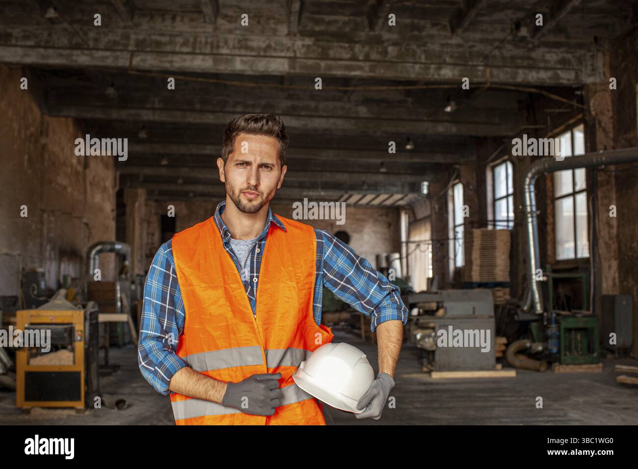 Porträt eines gutaussehenden männlichen jungen Fabrikarbeiters in orangefarbener Weste und Schutzhandschuhen, der einen weißen Schutzhelm in der Hand hält. Fabrikarbeiter nach h Stockfoto