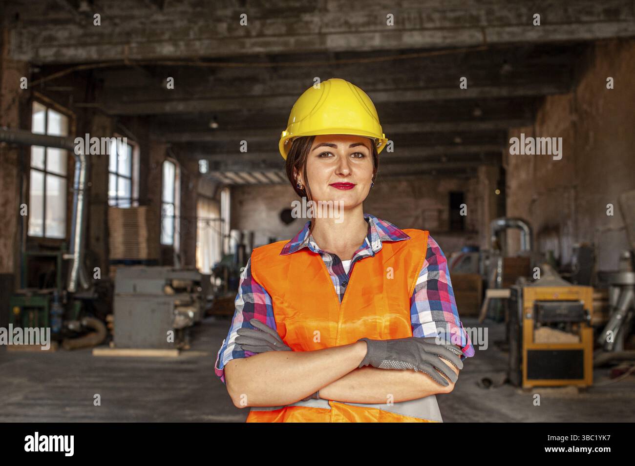 Nahporträt einer attraktiven jungen Arbeiterin in der Fabrik, trägt einen gelben Schutzhelm und graue Schutzhandschuhe mit verschränkten Armen und schaut auf die Stockfoto