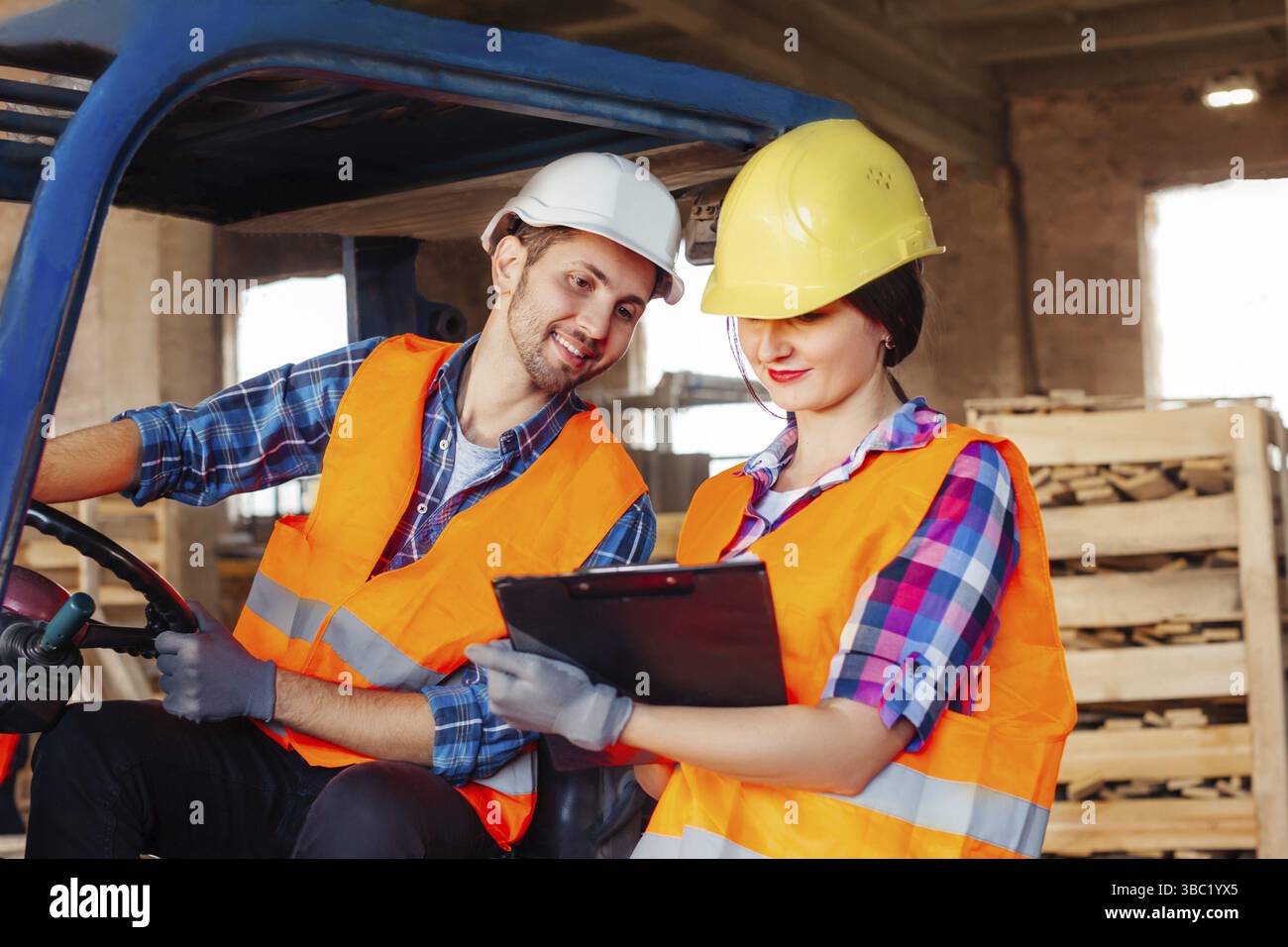 Ein Gabelstaplerfahrer sitzt in einem LKW, eine Fabrikarbeiterin steht in der Nähe, beide diskutieren Arbeitsprobleme. Lagerarbeiter in Hardhats und Orange Stockfoto