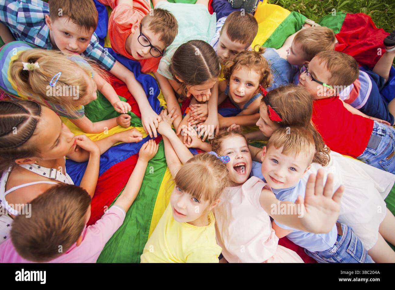 Draufsicht Kinder haben Spaß, legen sich auf eine bunte Matte und verbinden ihre Hände. Mehrere Kinder halten die Hände hoch, lächeln, schauen in die Kamera. Kind Stockfoto